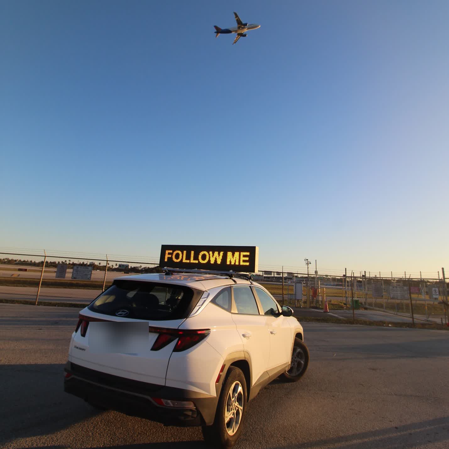 Truck-mounted high-visibility LED Follow Me signage vehicle on an airport apron with an aircraft departing overhead